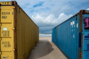 Blue and yellow shipping containers aligned on a sandy beach with the ocean and sky in the background.
